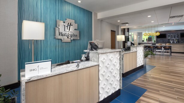 A reception area with a blue textured wall, a Holiday Inn sign, a reception desk with a phone and computer, and a bar area in the background. - Holiday Inn & Suites Arden - Asheville Airport by IHG (Arden)