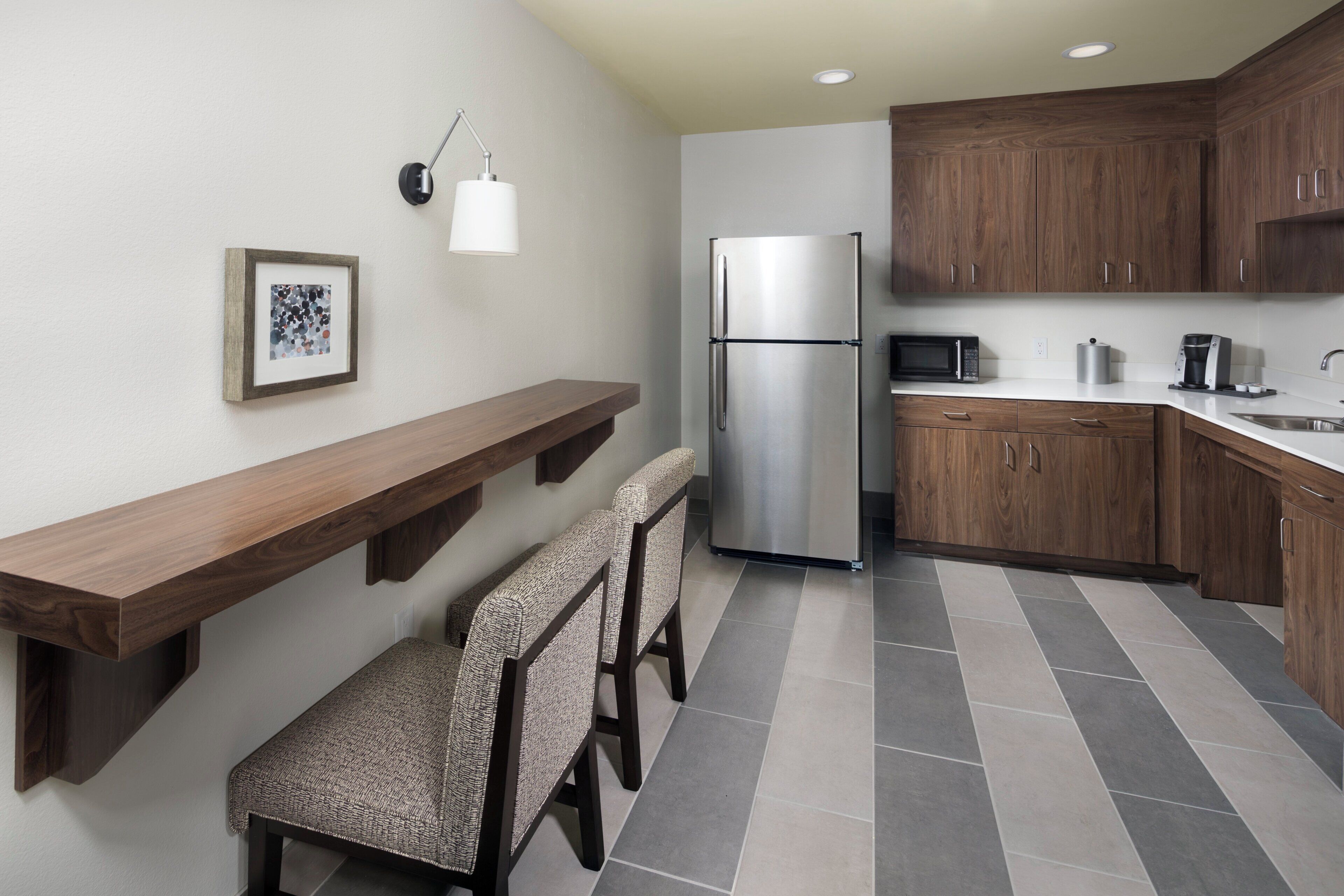 A modern kitchen with wooden cabinets, a stainless steel refrigerator, a microwave, and a coffee maker. There is a wooden bench with a woven seat and a wall-mounted light fixture.