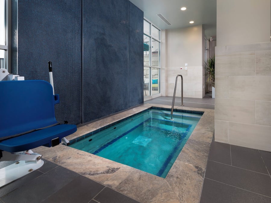 An indoor swimming pool with a blue chair, a metal ladder, and a tiled wall.