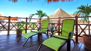 A wooden deck with green chairs and potted plants, a thatched roof, and a view of a pyramid structure. - Kaan 302 by NAS (Playa del Carmen)