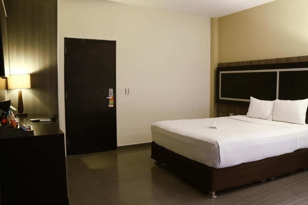 A hotel room with a large bed, a desk with a lamp, and a door.