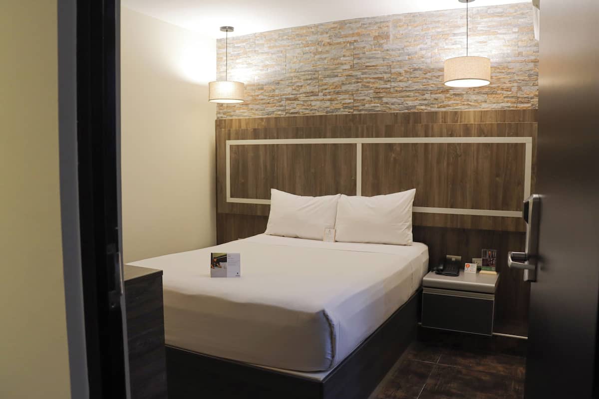 A modern hotel room with a neatly made bed, a bedside table with a phone and a lamp, and a stone-textured wall behind the bed.