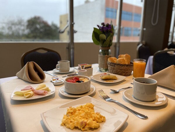 A breakfast table with plates of eggs, ham, and pastries, a vase with flowers, and a basket of bread. - Rado Hotel - Santa Maria (Trujillo)