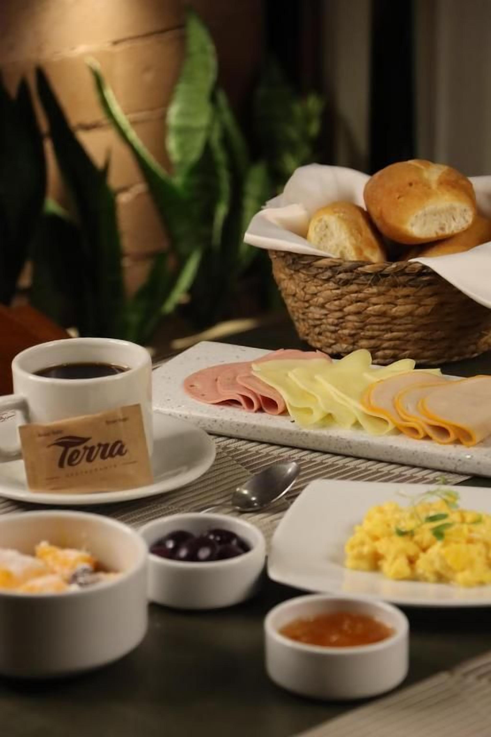 A breakfast setting with bread, ham, cheese, and eggs on a tray, a cup of coffee, and small bowls of olives and jam.
