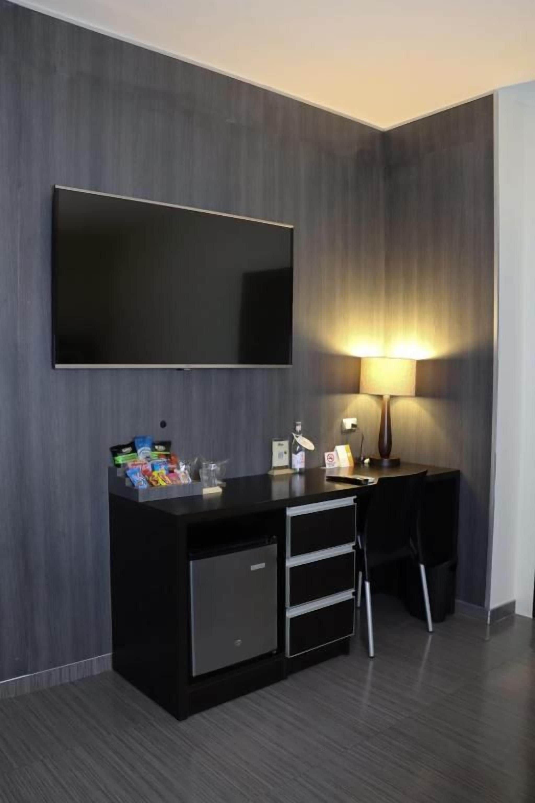 A modern hotel room with a flat-screen TV mounted on the wall, a desk with a lamp, and a small minibar.