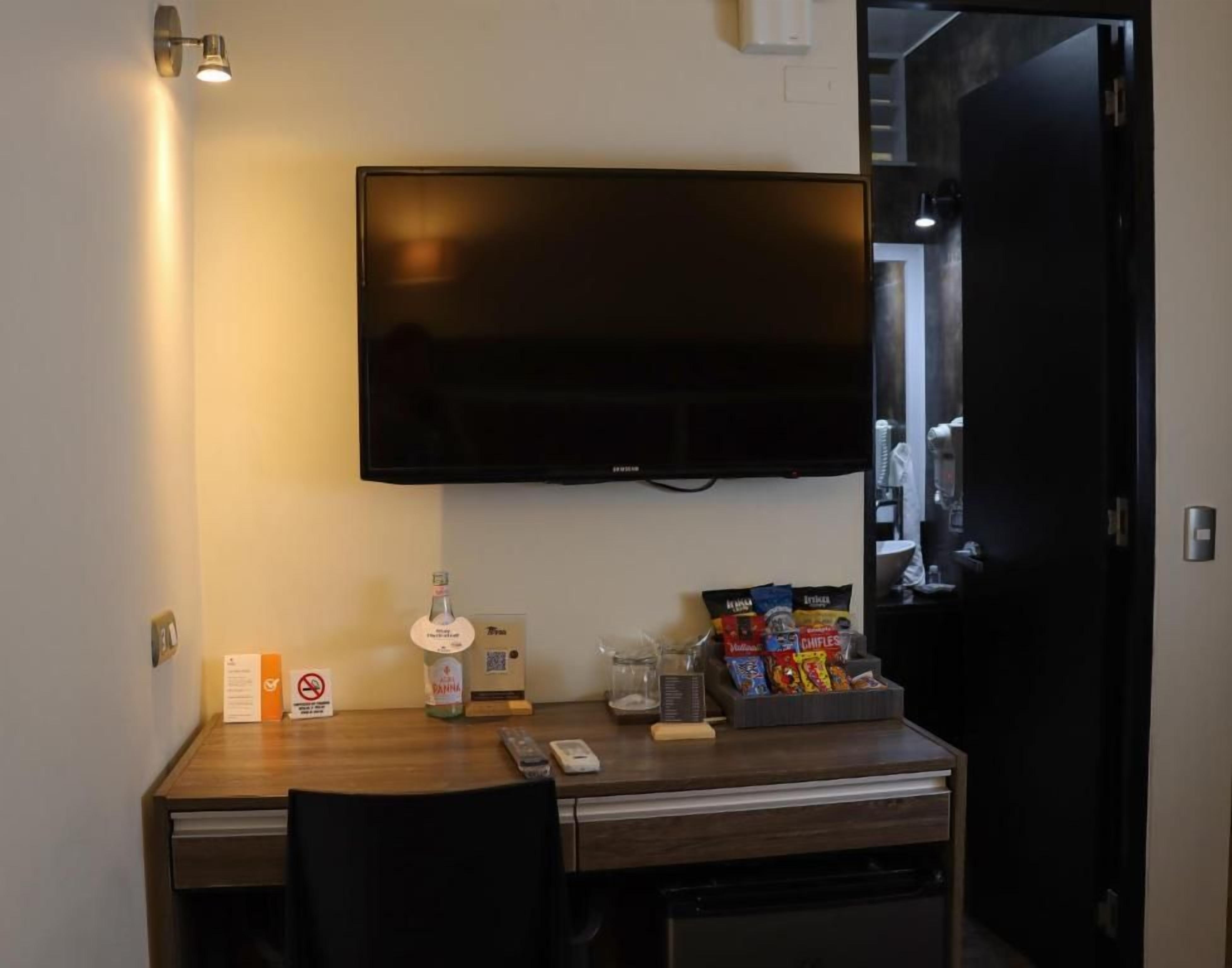 Ein Hotelzimmer mit einem an der Wand befestigten Flachbildfernseher, einem Holztisch mit einer Wasserflasche, Snacks und einem Glas.