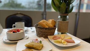 Desayuno con croissant, tabla de jamón y queso, frutero y cesta de pan.