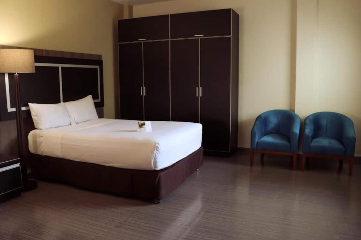 A hotel room with a large bed, a wardrobe, two blue chairs, and a lamp.