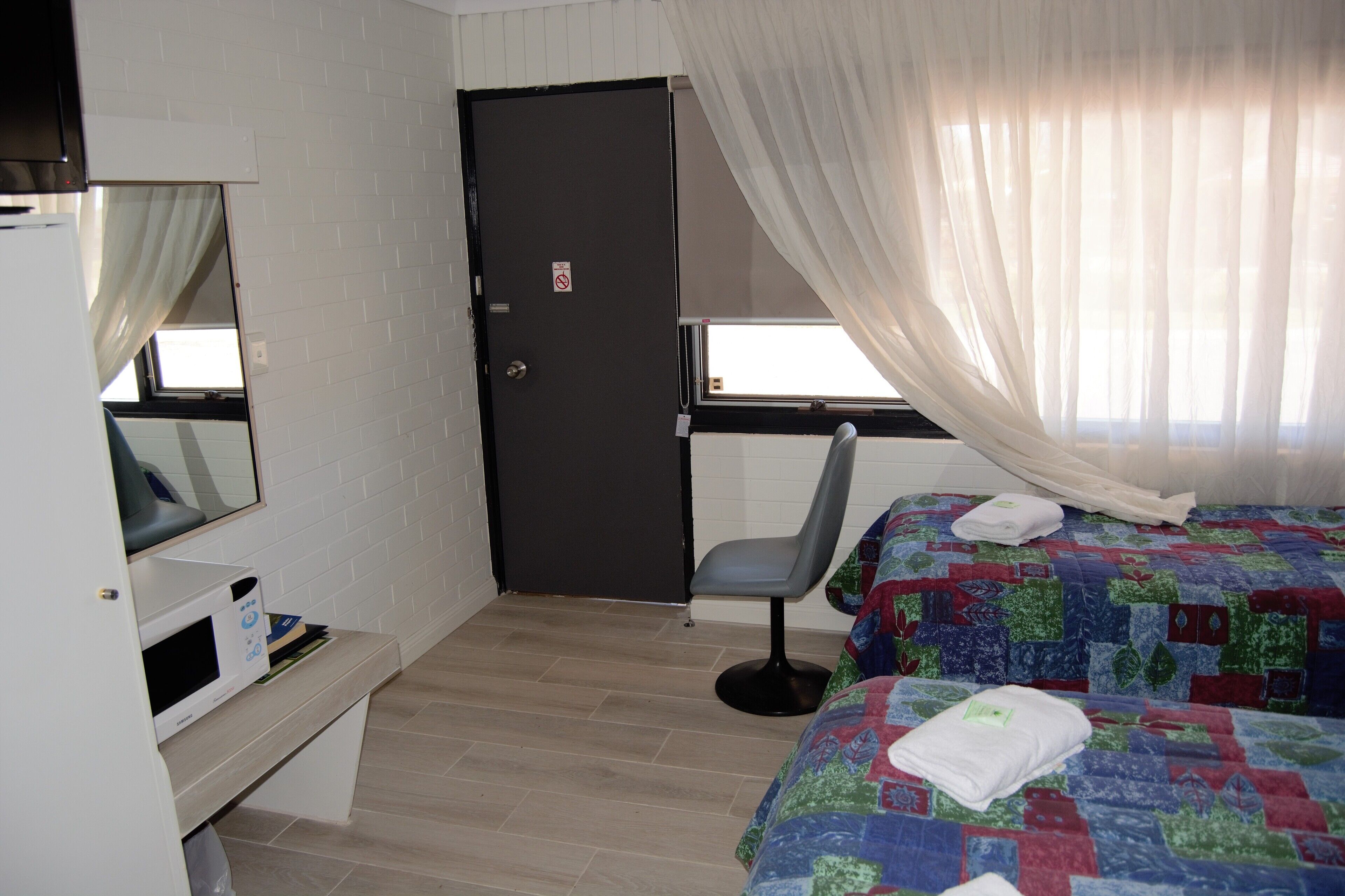 A bedroom with a bed, a chair, a desk with a computer, a door, and a window with curtains.