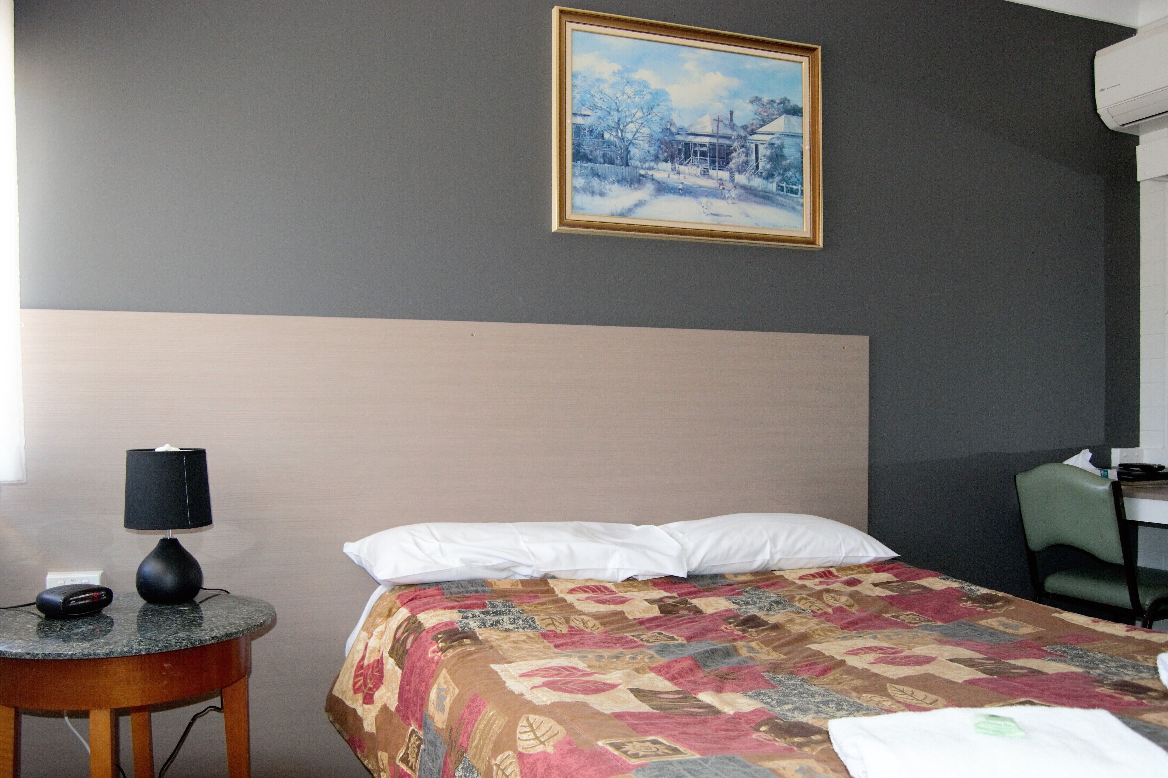 Una habitación de hotel con una cama, una mesa redonda con lámpara, un cuadro en la pared y una silla.