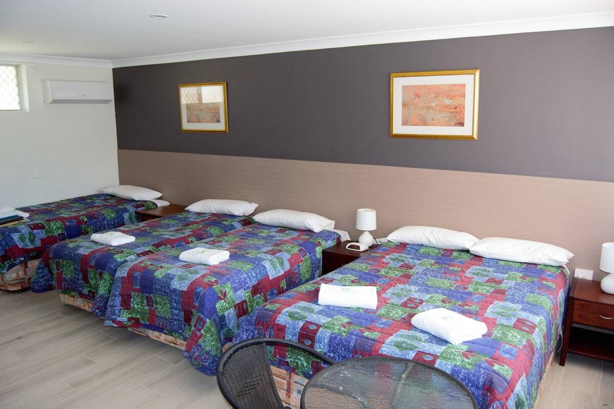 A hotel room with three beds, each with a colorful quilt and white pillows. There is a small table with a chair, a bedside table with a lamp, and a framed picture on the wall.