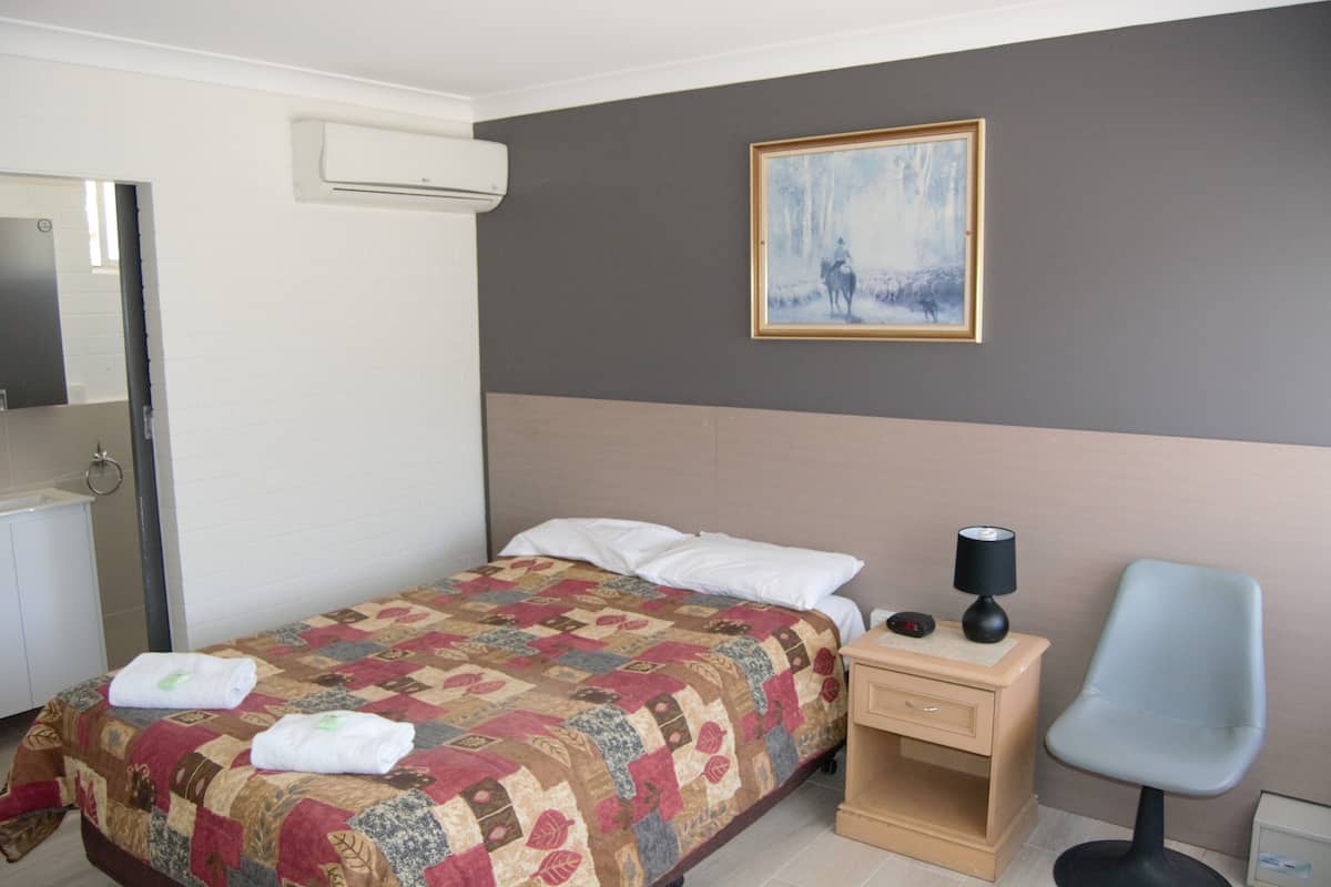A hotel room with a bed, a nightstand, a chair, a painting on the wall, and a bathroom with a mirror and sink.