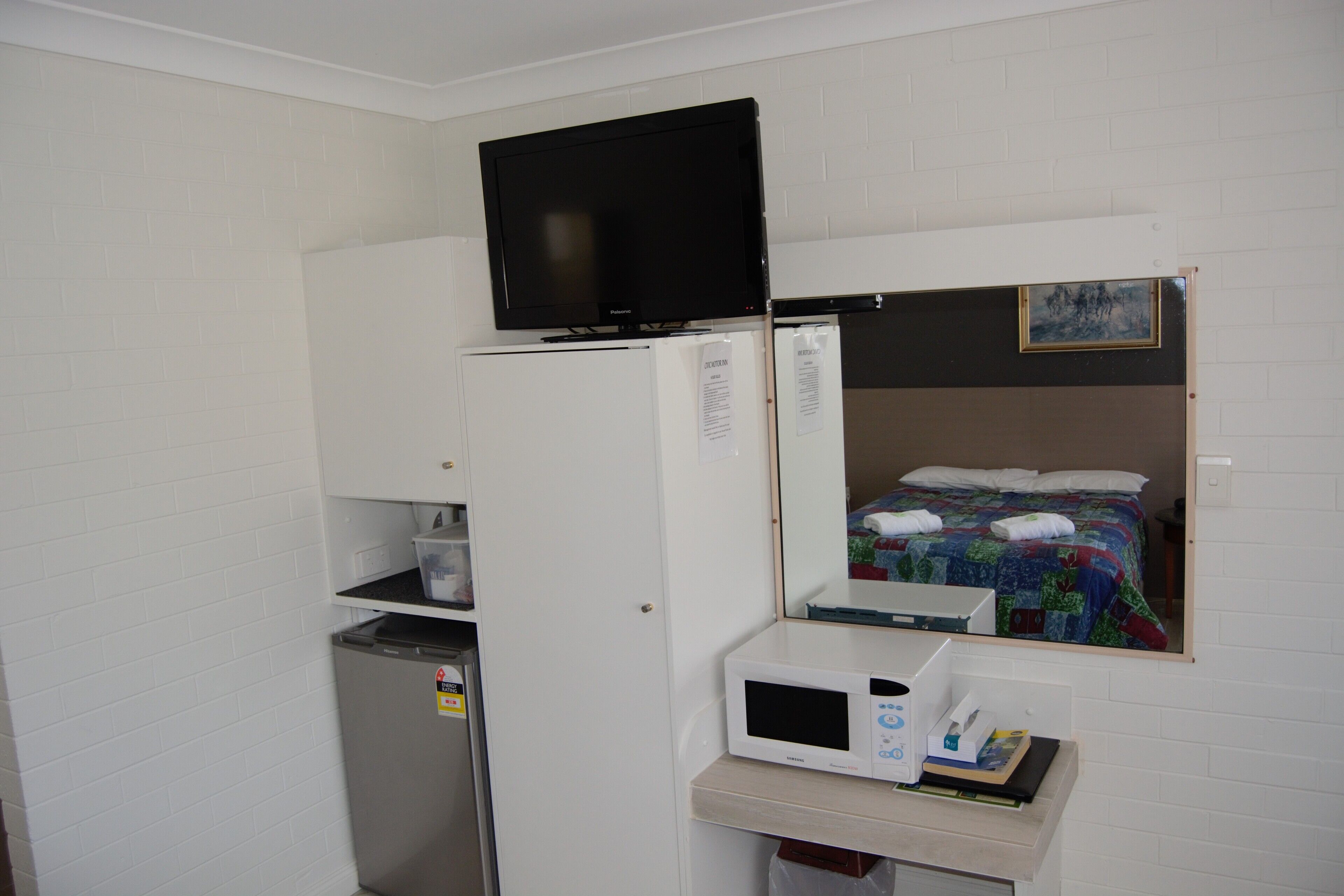 A compact room with a bed, a television on top of a white cabinet, a microwave, and a small refrigerator.