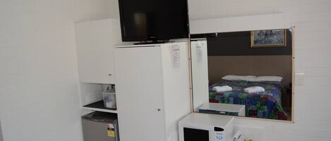 A compact room with a bed, a television on top of a white cabinet, a microwave, and a small refrigerator.