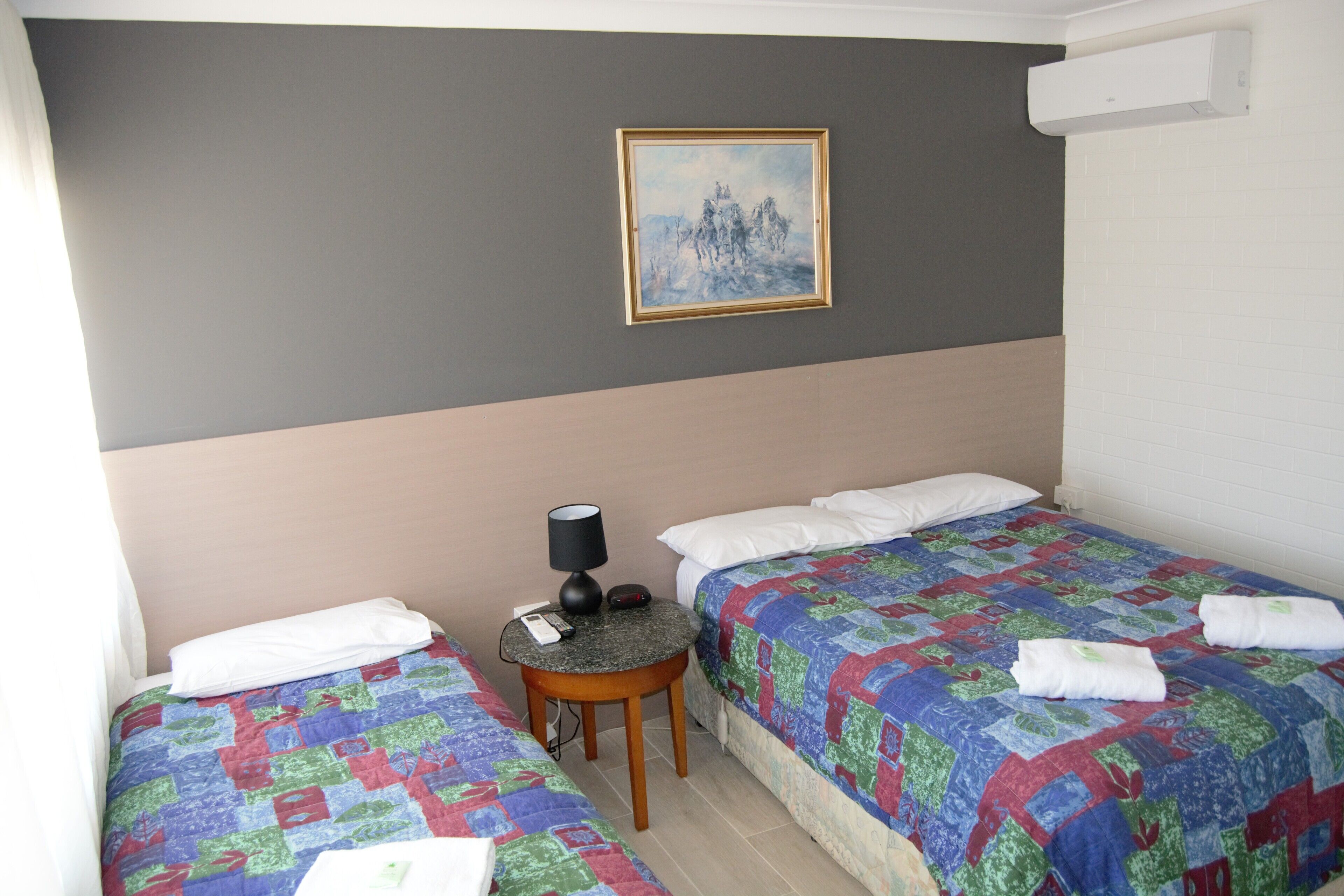 Ein Hotelzimmer mit zwei Betten, einem kleinen Tisch mit einer Lampe, einem Bild an der Wand und einer Klimaanlage.
