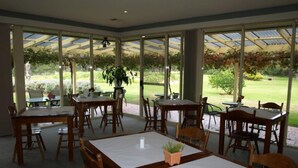 Um restaurante com várias mesas e cadeiras, grandes janelas e vista para um jardim.