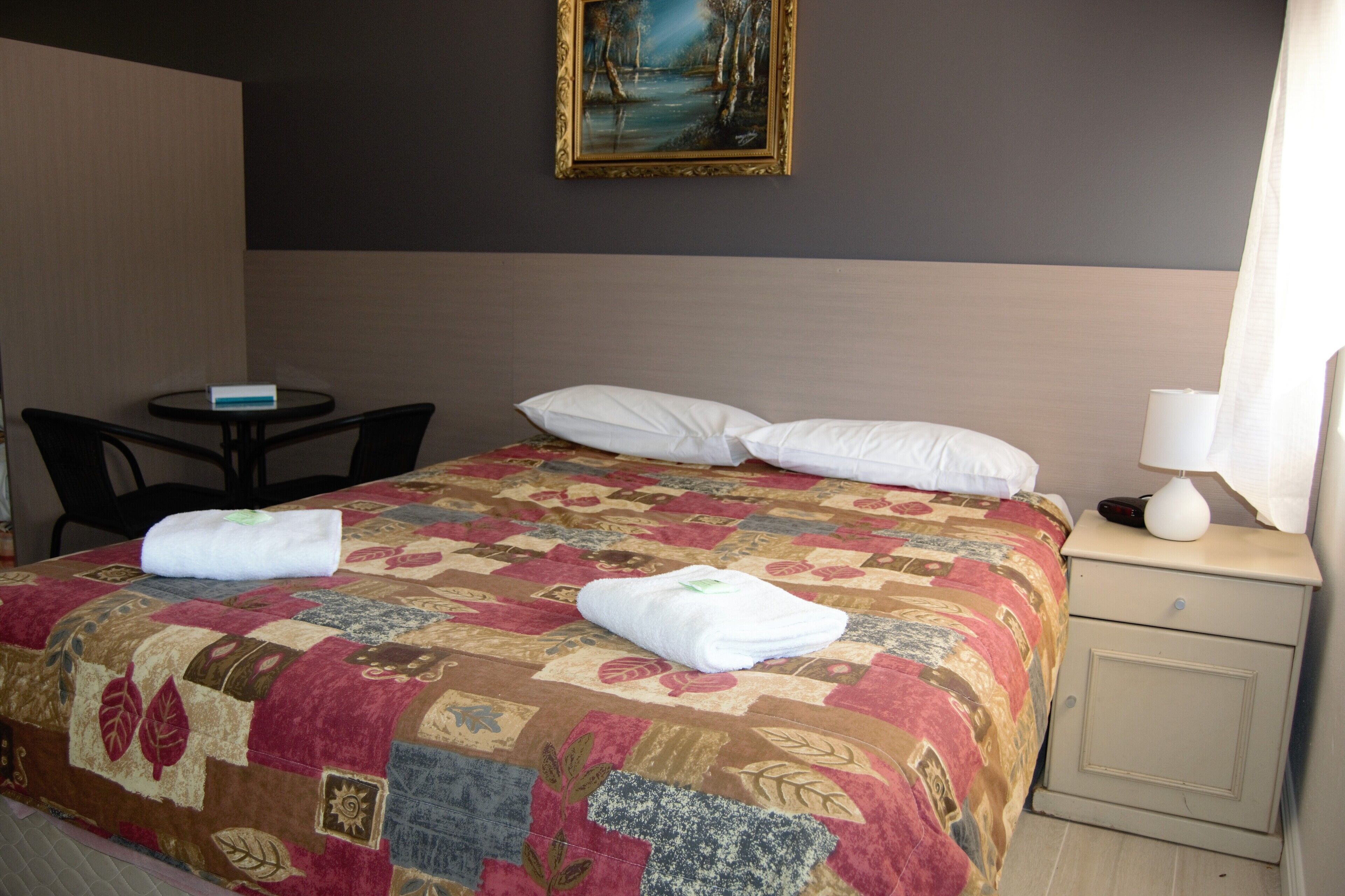 A hotel room with a bed, a small table, a chair, a nightstand, and a painting on the wall.