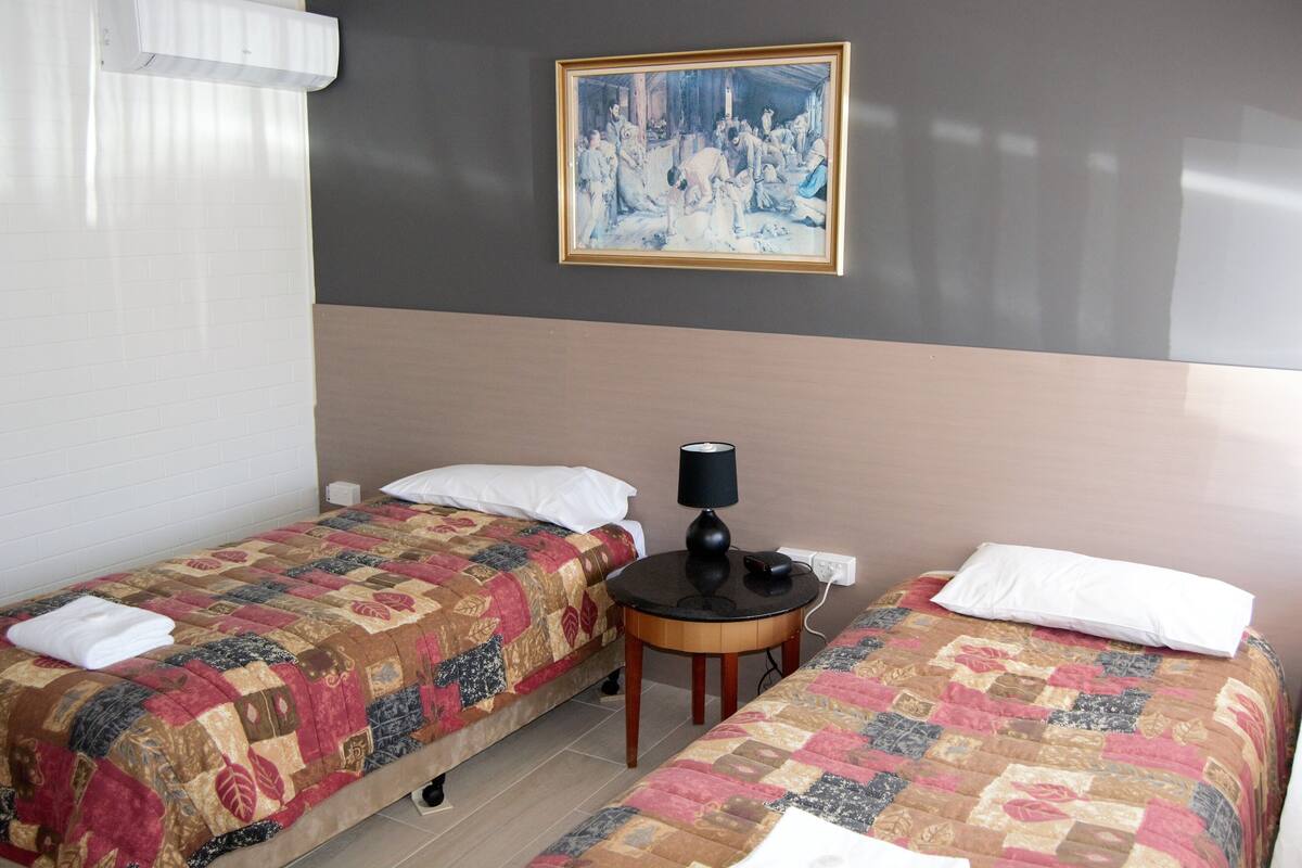A hotel room with two beds, a painting on the wall, a small table with a lamp, and a window with blinds.
