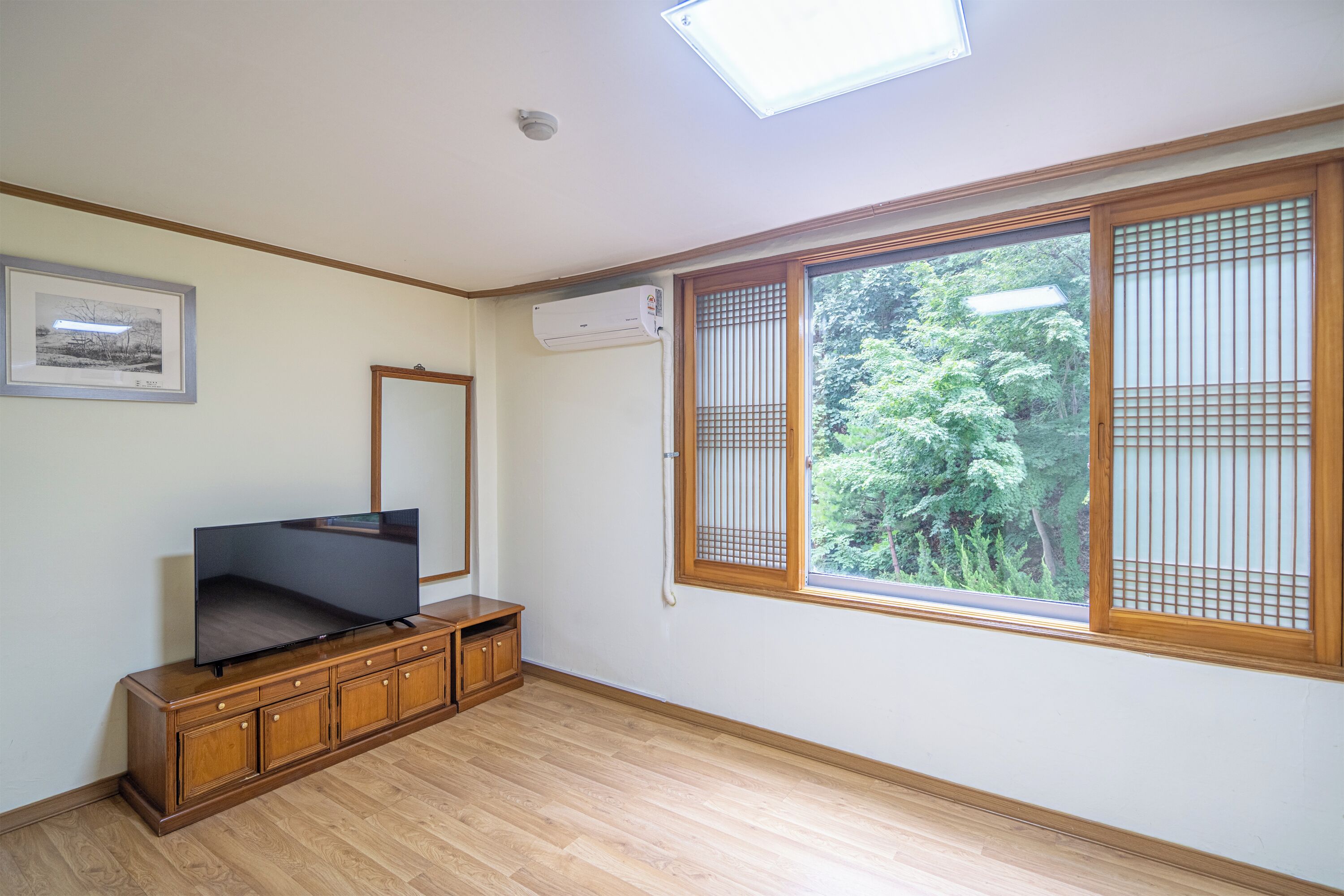 A room with a wooden cabinet, a flat-screen TV, a ceiling fan, and a large window with a view of greenery.