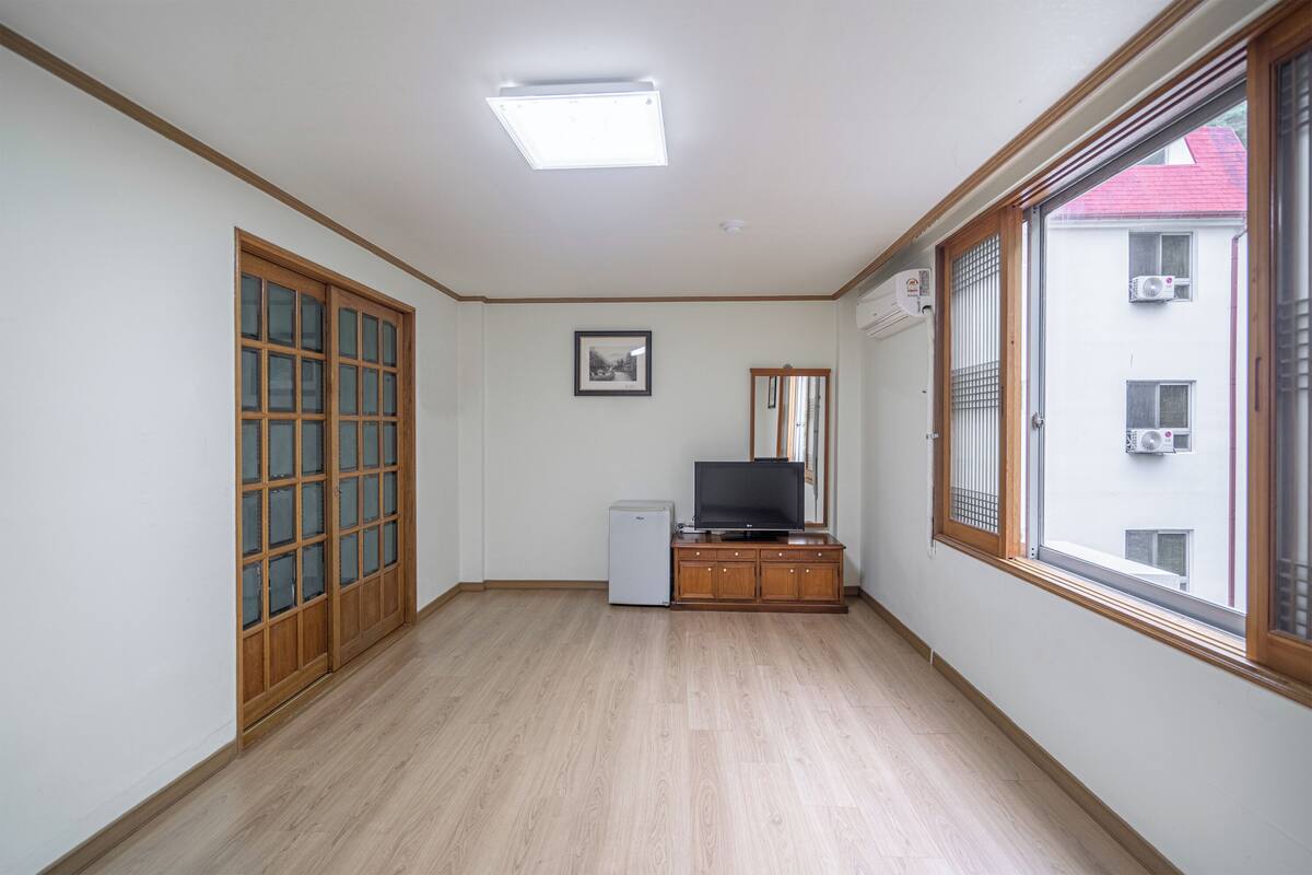 Ein Zimmer mit Holzboden, einem Fernseher auf einem Schrank, einem kleinen Kühlschrank und einem Fenster mit Blick auf Gebäude.