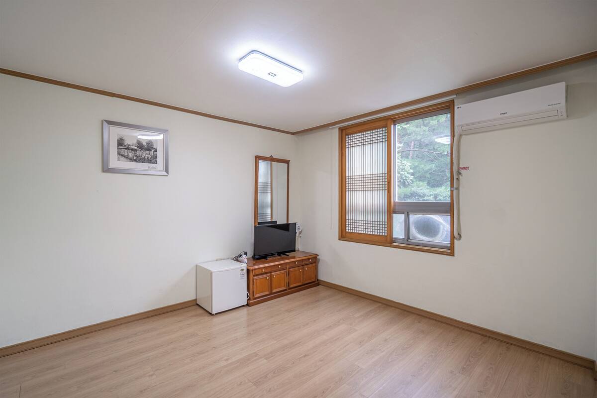Ein Zimmer mit Holzboden, einem kleinen weißen Kühlschrank, einem Fernseher auf einer Holzkommode, einem gerahmten Bild an der Wand und einem Fenster mit Jalousien.
