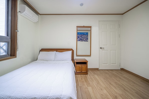 Un dormitorio con una cama, una mesita de noche, piso de madera, una puerta blanca y una ventana con aire acondicionado.