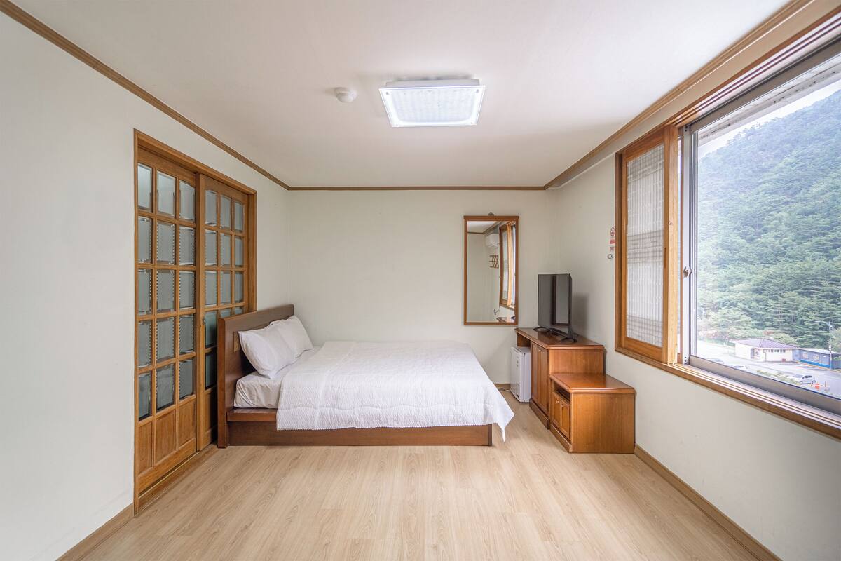 Ein Schlafzimmer mit Bett, Fernseher, Holzschrank, Fenster mit Blick auf einen Wald und Deckenleuchte.
