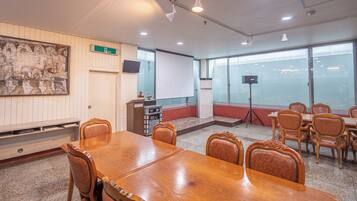 Un salón de conferencias con una mesa y sillas de madera, una pizarra, un proyector y un amplio ventanal con persianas.
