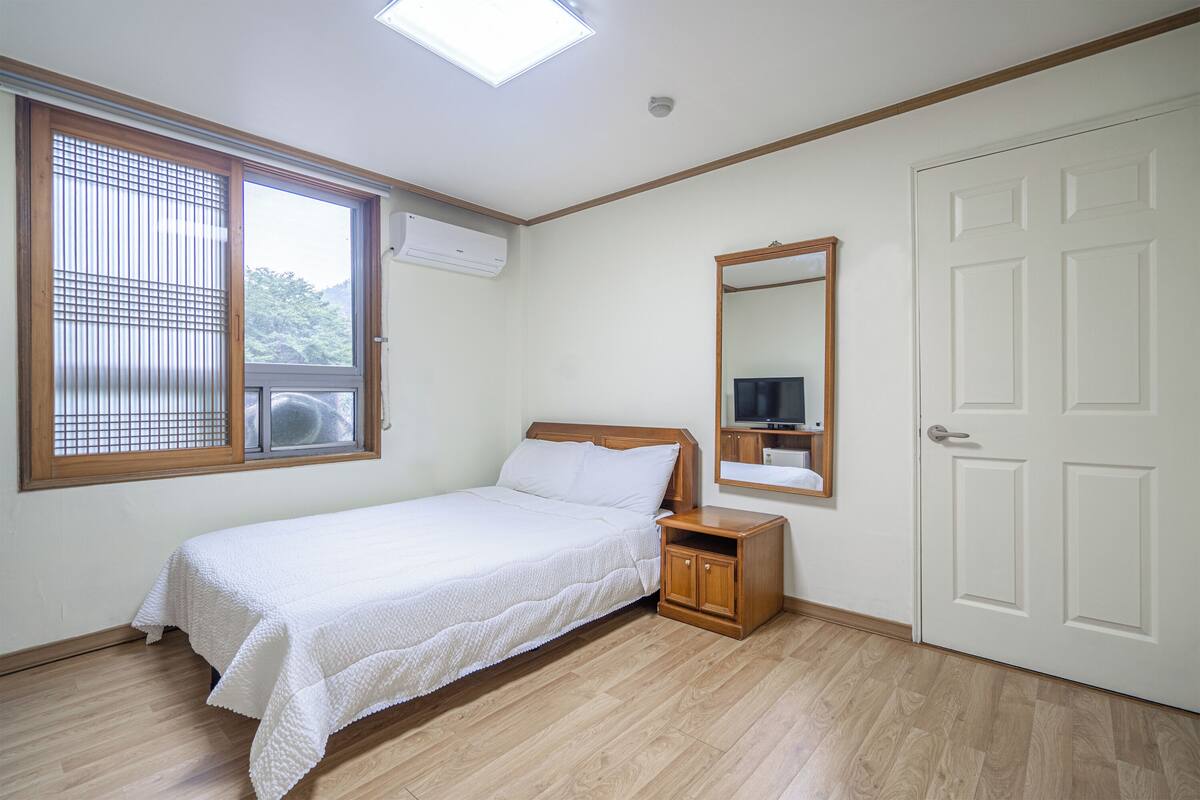 Ein Schlafzimmer mit Bett, Nachttisch, Spiegel, Kleiderschrank und einem Fenster mit Jalousien.