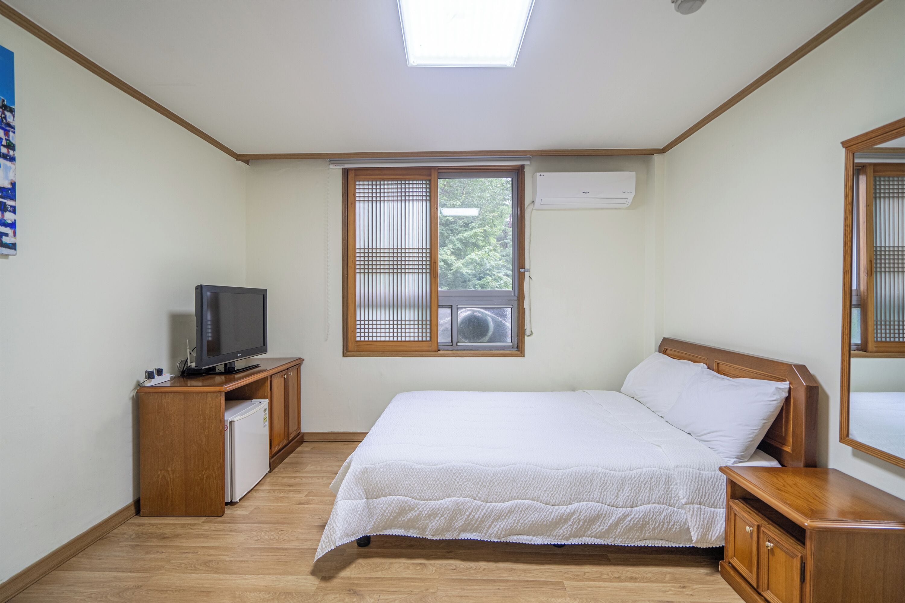 A bedroom with a bed, a television on a wooden stand, a small refrigerator, and a window with blinds.