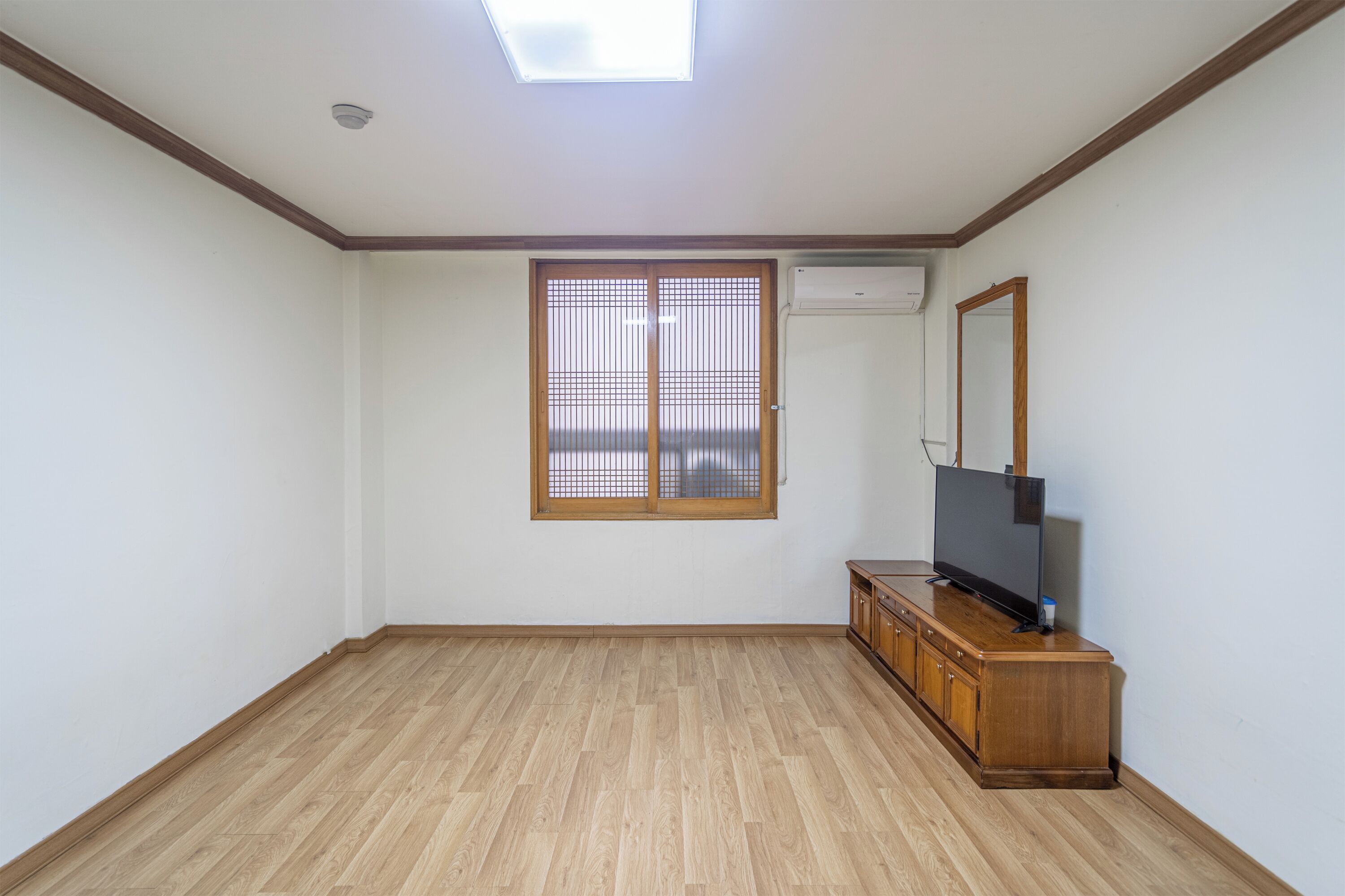 A room with a wooden floor, a large window with blinds, a television on a wooden cabinet, and an air conditioning unit on the wall.