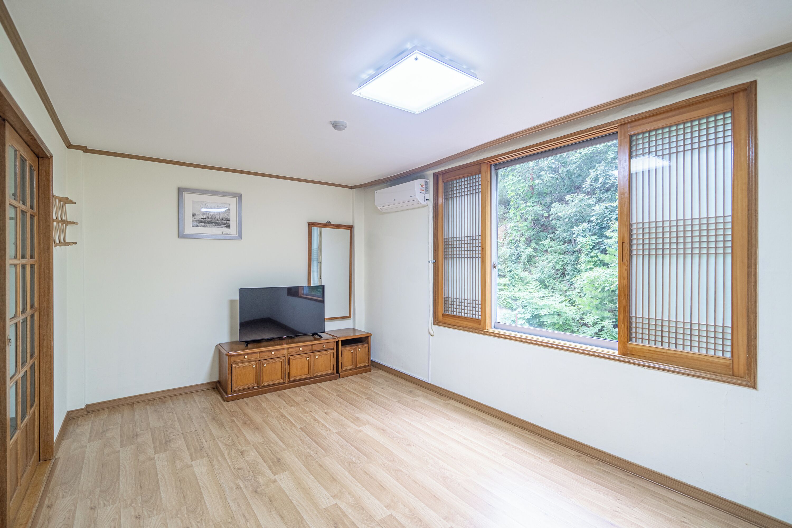 A room with a large window, wooden flooring, a flat-screen TV, and a small cabinet.