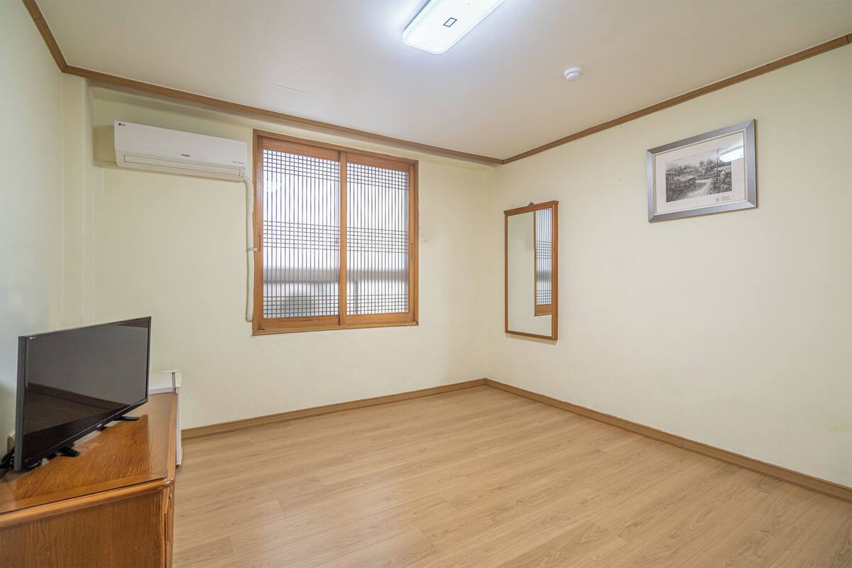 Ein Zimmer mit Holzboden, einem Flachbildfernseher auf einer Holzkassette, einem Fenster mit Holzjalousien und einem gerahmten Bild an der Wand.