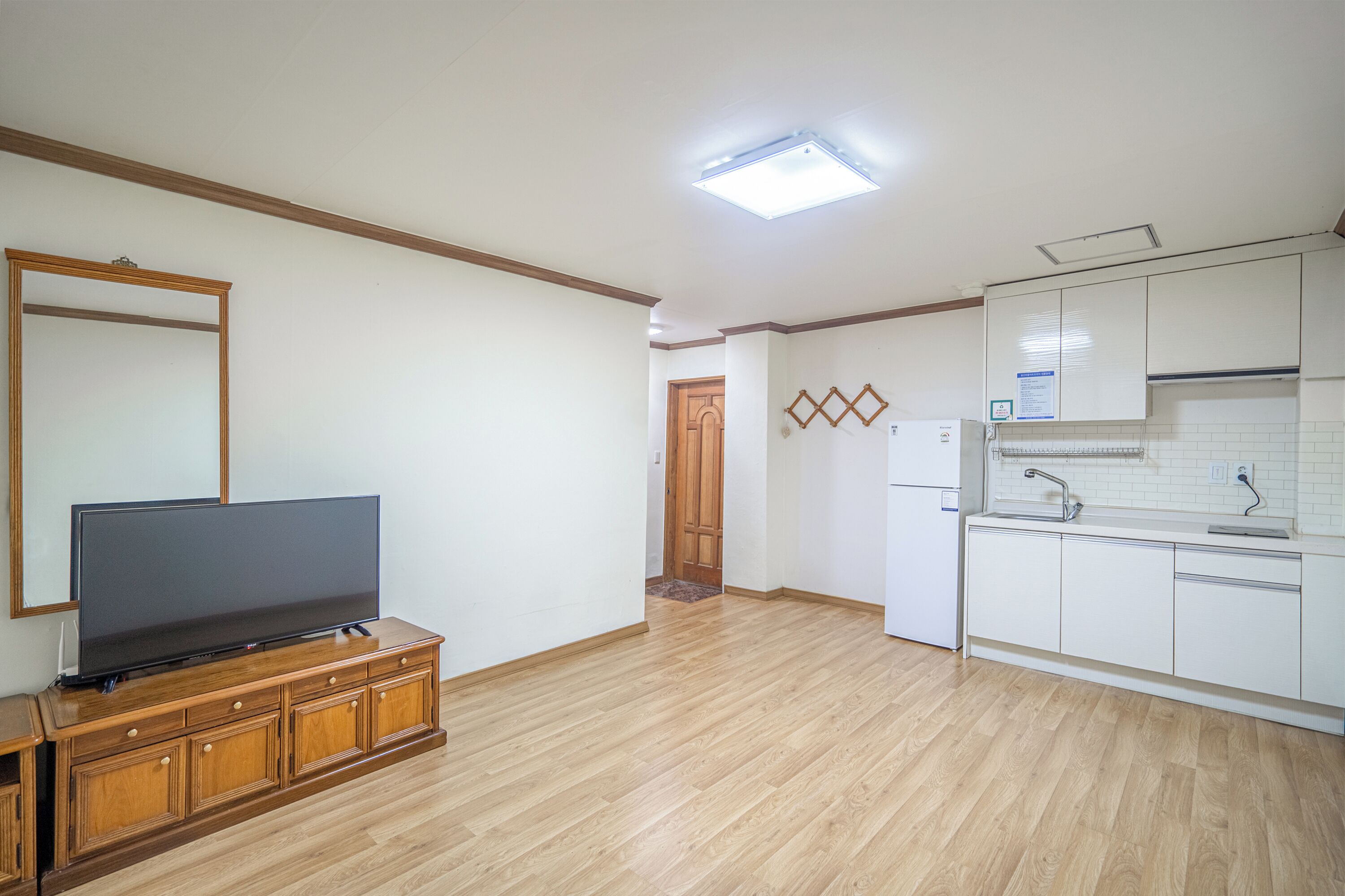 Amplia sala de estar con televisor de pantalla plana sobre un mueble de madera, cocina con armarios blancos y horno empotrado, y puerta de madera que da a otra habitación.