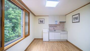 Cocina pequeña con refrigerador, lavamanos y piso de madera. Se ve un ventanal con vistas a árboles y una lámpara en el techo.
