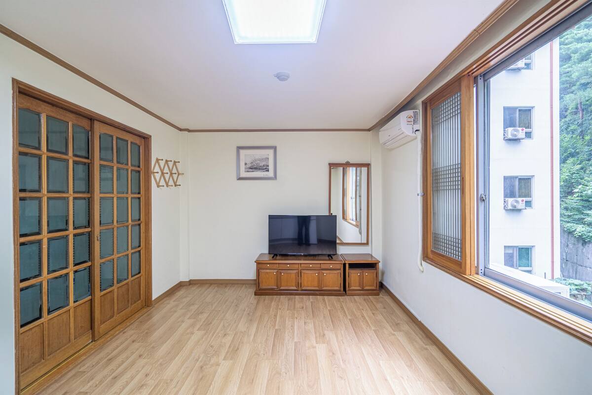Ein Zimmer mit einem Holzschrank, einem Fernseher, einem gerahmten Bild und einem Fenster mit Blick auf Gebäude.