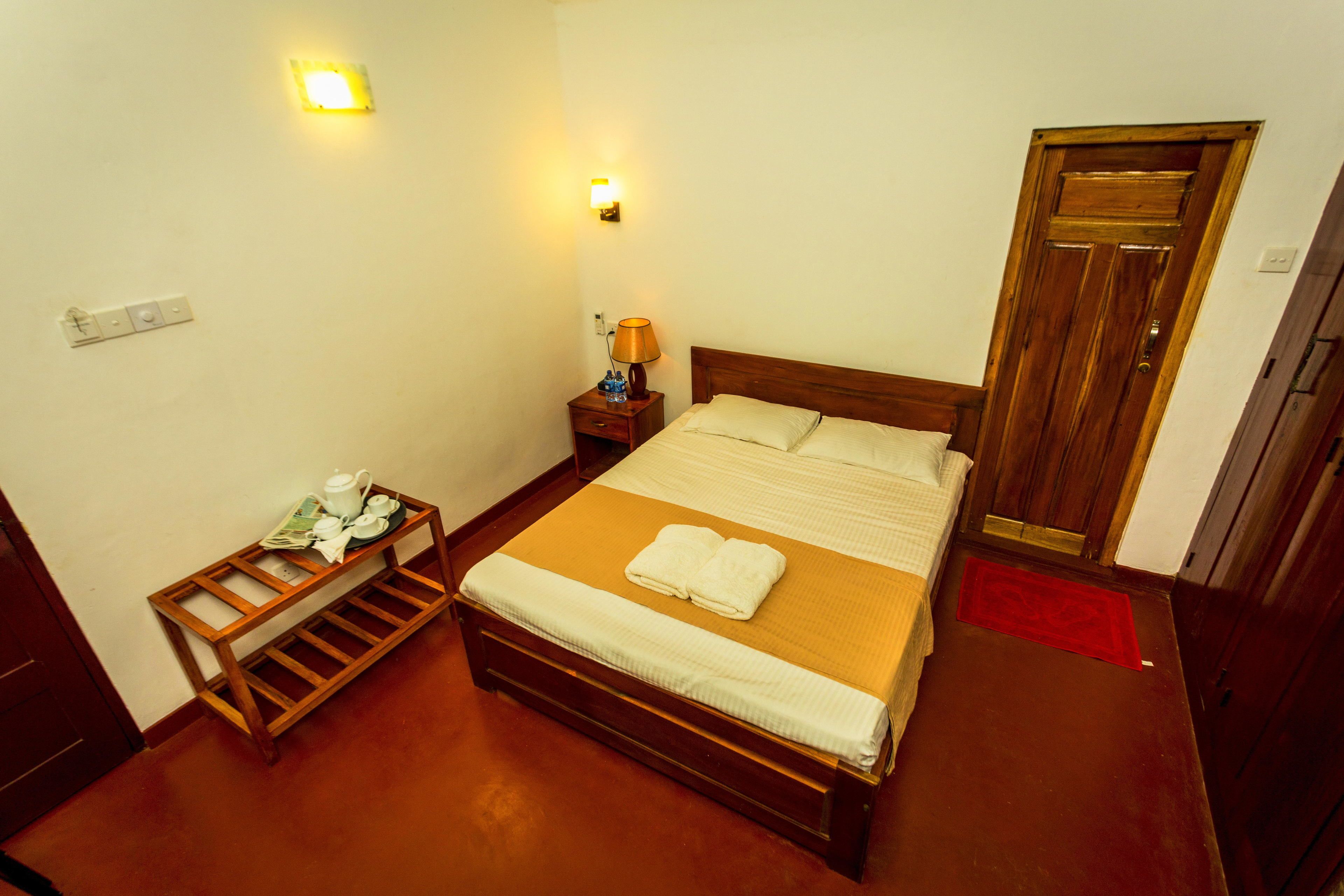 A bedroom with a wooden bed, a bedside table with a lamp, a small shelf, and a door.
