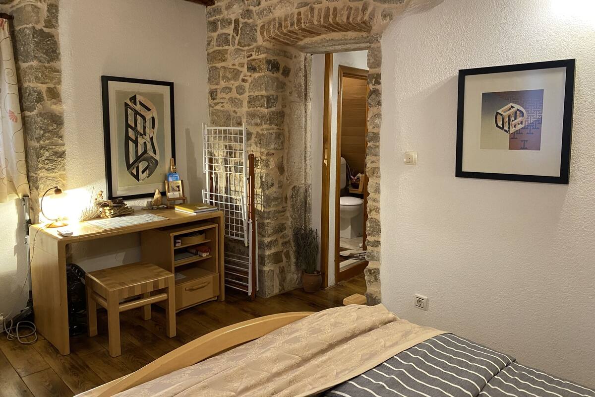 Un dormitori amb un llit de fusta, un escriptori amb una cadira, una paret de pedra i una porta que dóna a un bany.