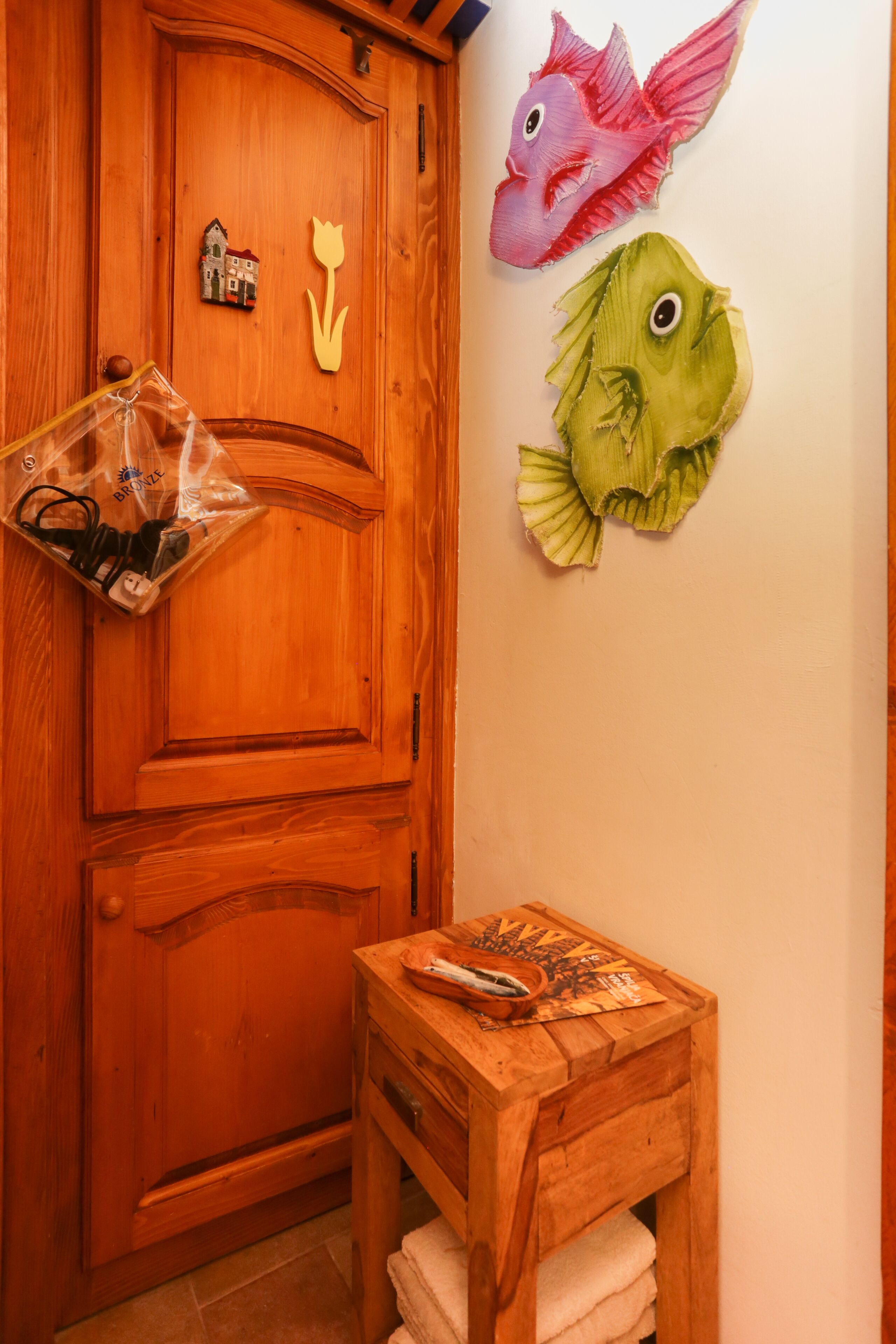 Een houten deur met een plastic zak er aan, een klein houten tafeltje met een handdoek, en twee vissenornamenten aan de muur.
