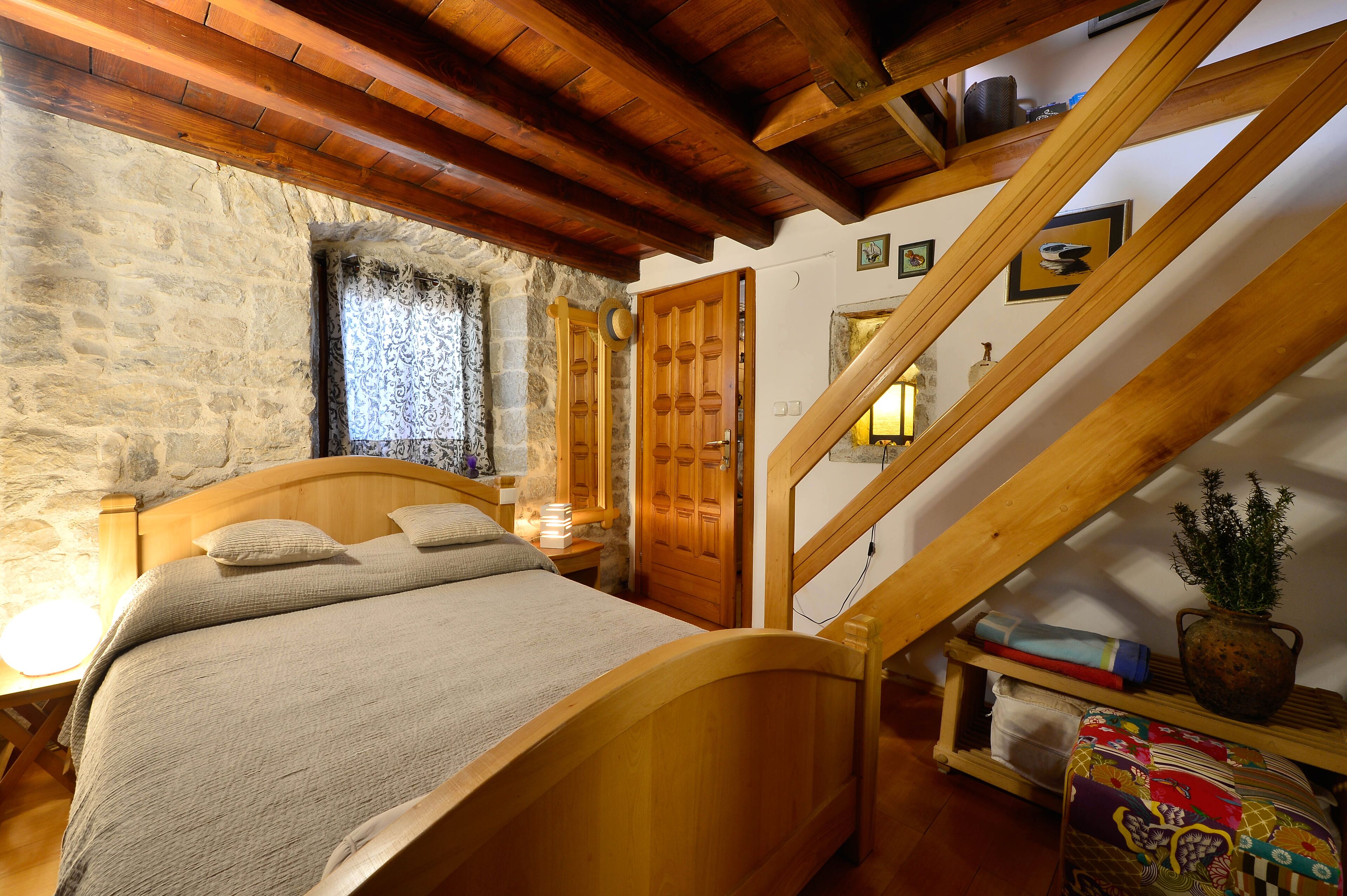 Um quarto aconchegante com uma cama de madeira, parede de pedra, escada de madeira e uma pequena mesa com luminária.