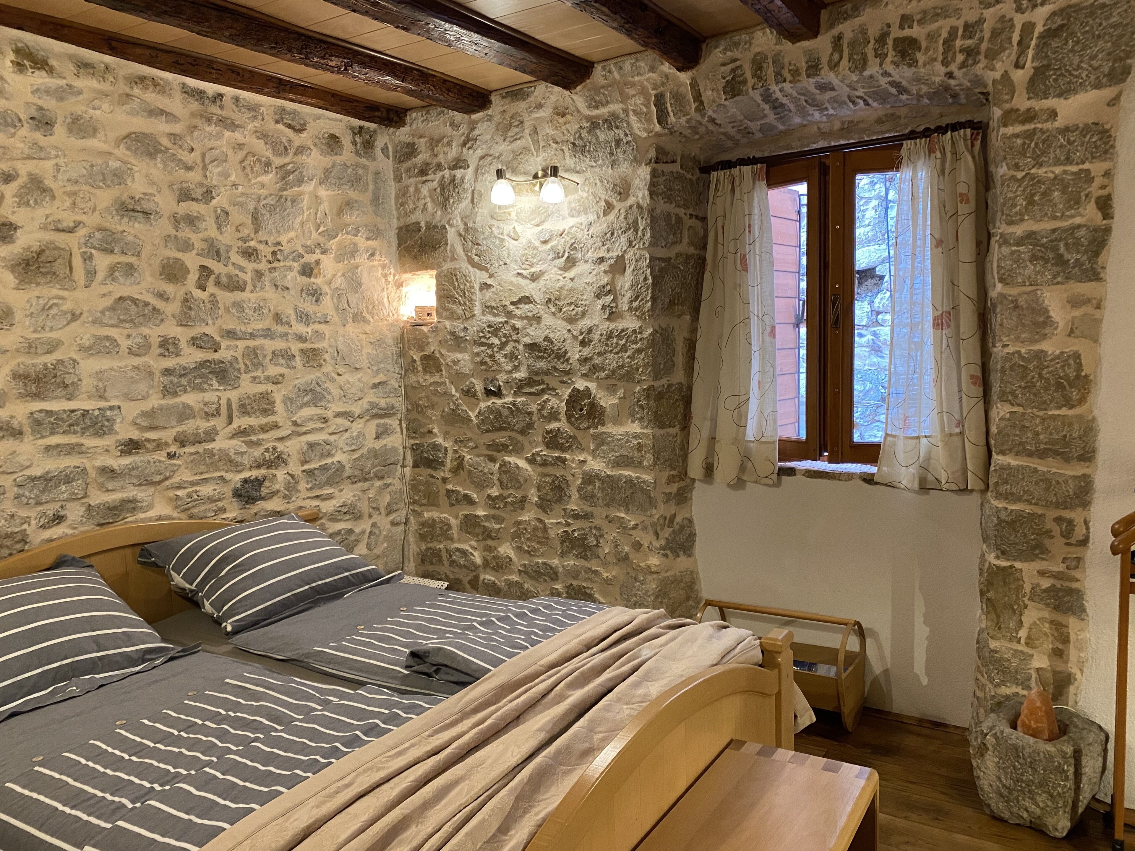 Ein Schlafzimmer mit Steinwänden, einem hölzernen Bett, einem kleinen Tisch und einem Fenster mit Vorhängen.
