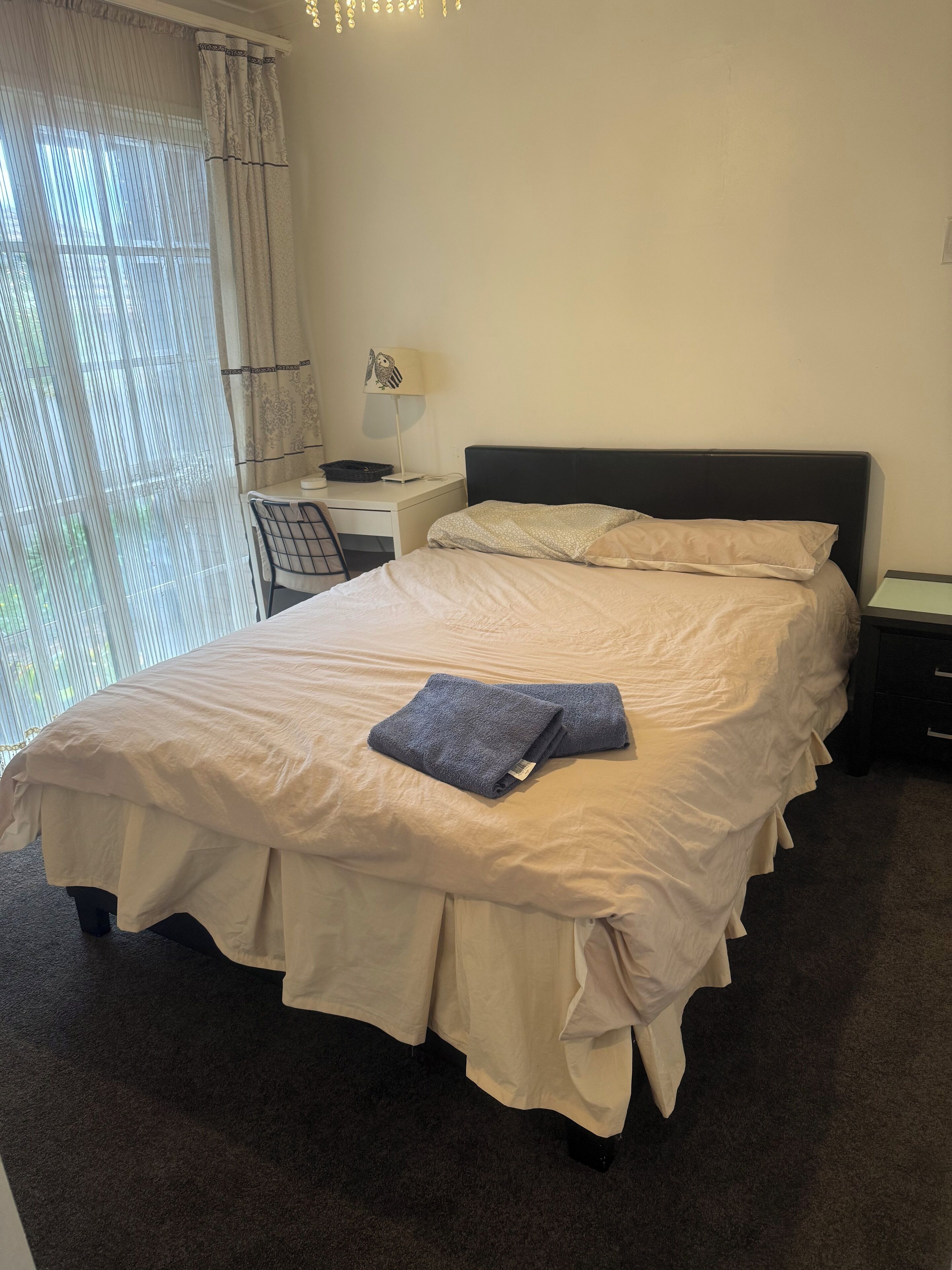 Una cama bien hecha con edredón y almohadas, una mesita de noche con lámpara, una silla y un escritorio.
