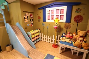 A children's playroom with a slide, storage units, a white picket fence, and a bench with teddy bears.