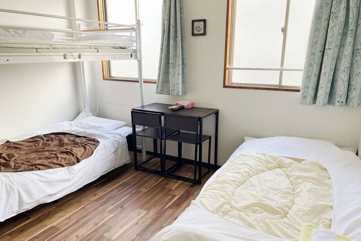 A dorm-style room with two beds, a small desk, and a window with curtains.
