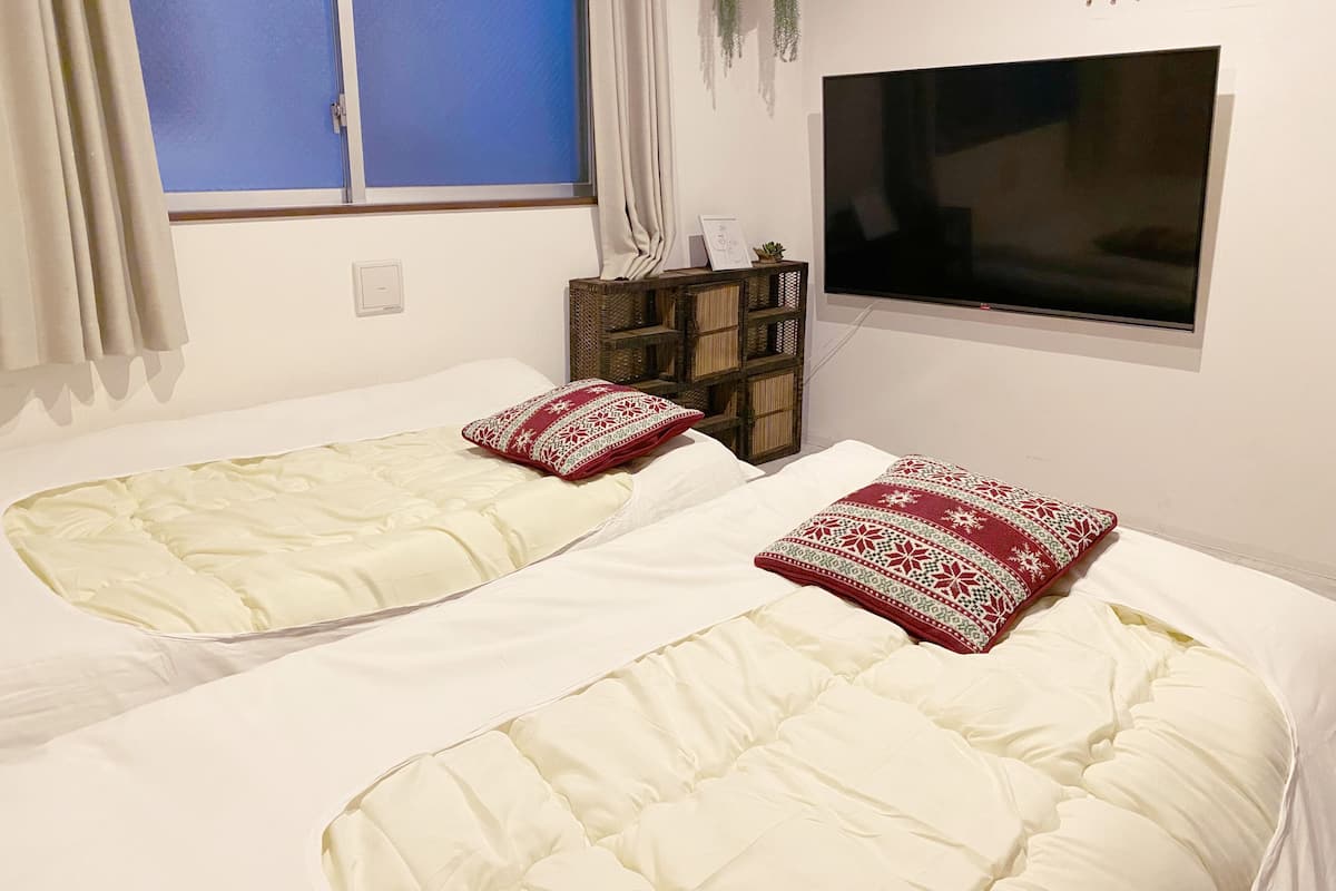 Two single beds with white bedding and red patterned pillows, a TV mounted on the wall, and a small wooden shelf with decorative items.