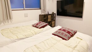 Dos camas individuales con ropa blanca y almohadas rojas estampadas, un televisor montado en la pared y un pequeño estante de madera con adornos.
