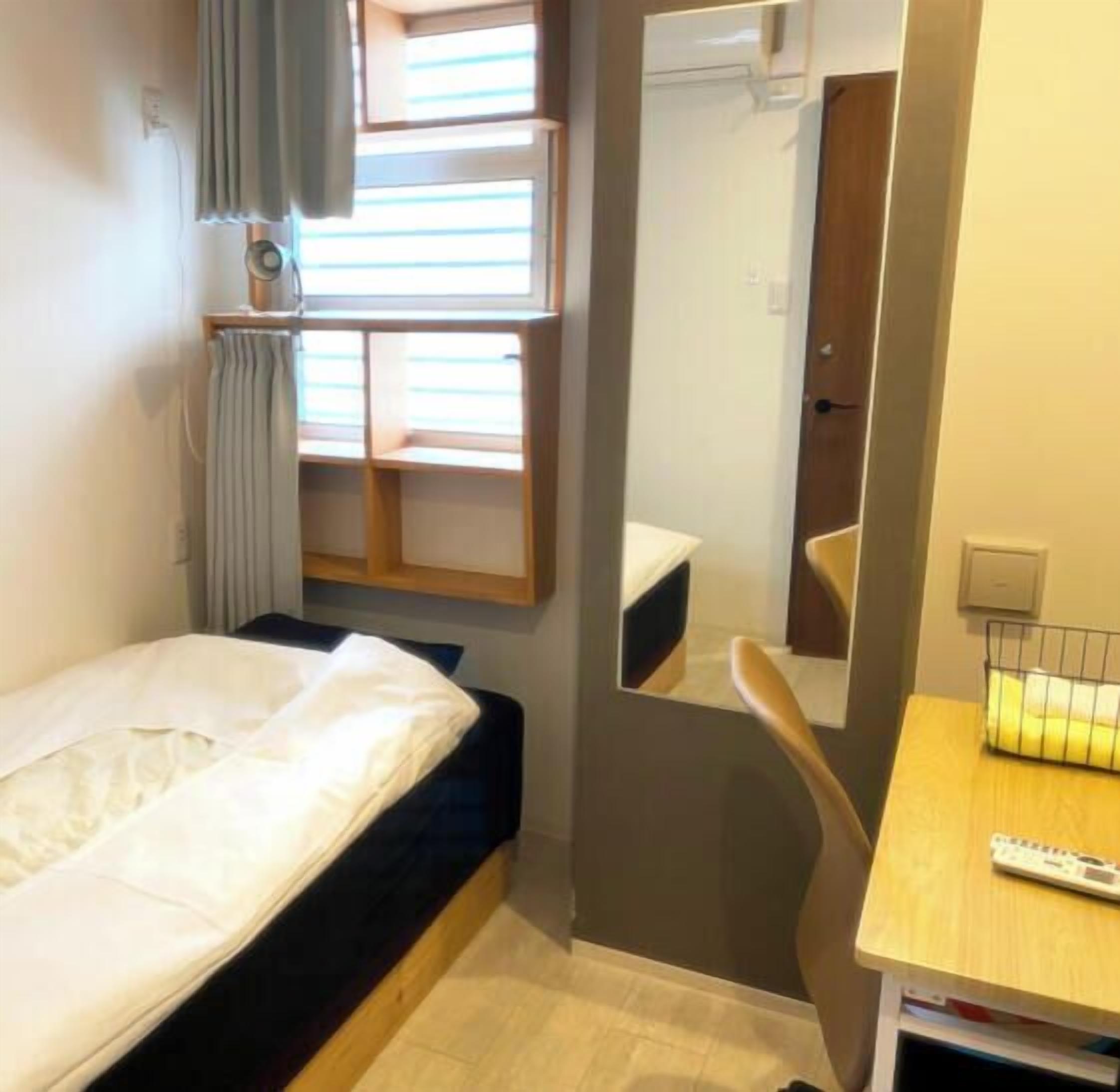 Ein kleines Hotelzimmer mit einem Bett, einem Spiegel, einem Schreibtisch mit Fernbedienung und einem Fenster mit Jalousien.