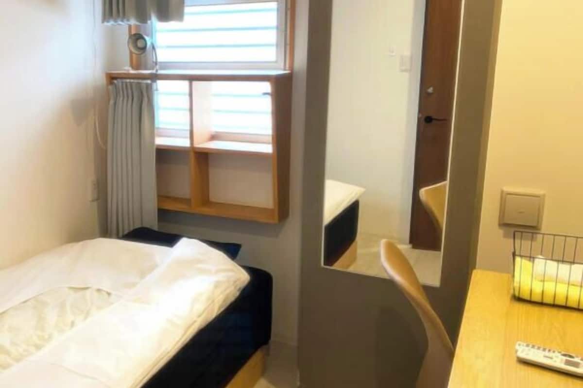 A small hotel room with a bed, a mirror, a desk with a remote control, and a window with blinds.