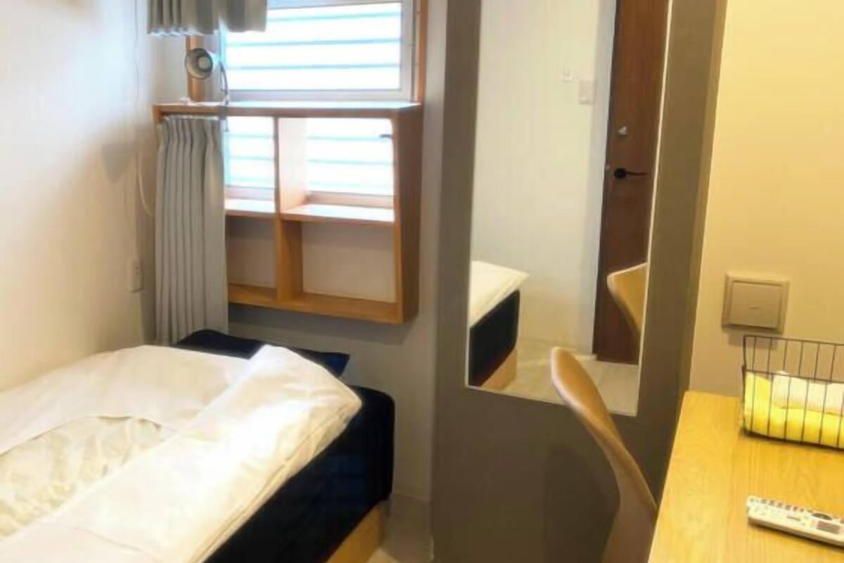 Ein kleines Hotelzimmer mit einem Bett, einem Spiegel, einem Schreibtisch mit Fernbedienung und einem Fenster mit Jalousien.