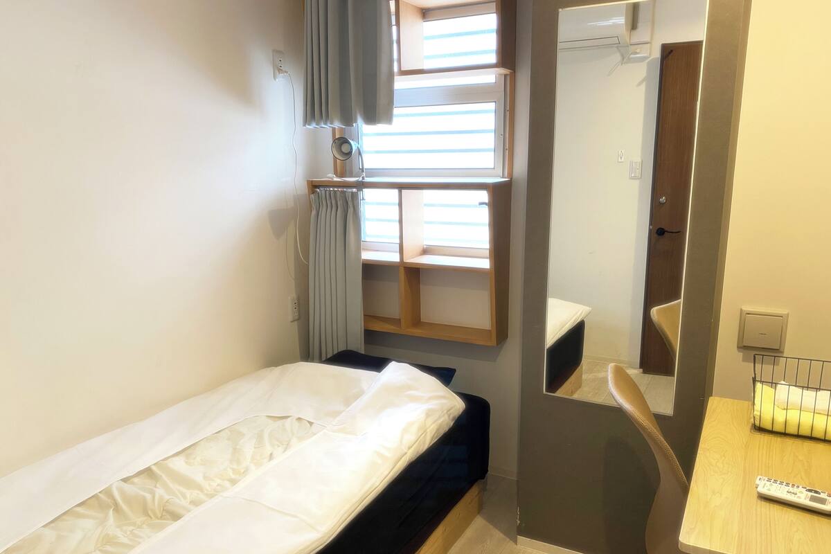A compact hotel room with a single bed, a small desk with a chair, a mirror, and a window with curtains.